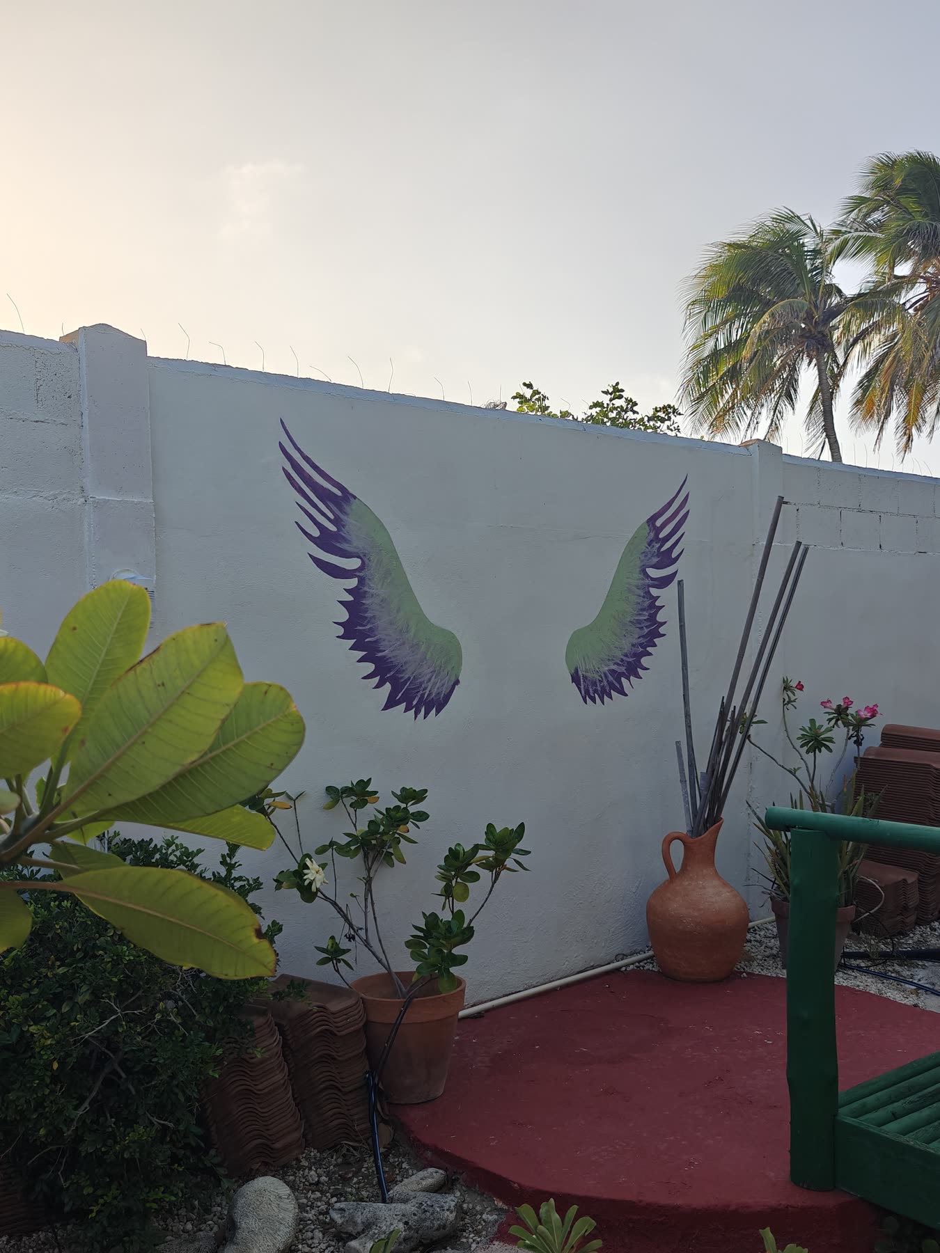 A pair of angel wings painted in purple and green on the white garden wall, above a round red patio