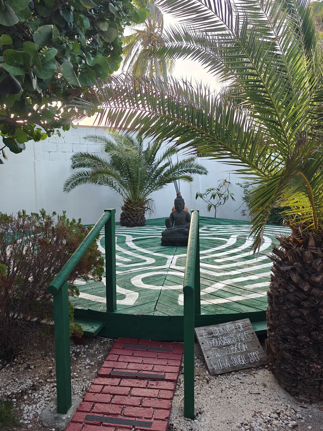 A seated Buddha at the centre of the raised meditation deck, its green boards painted with a white labyrinth, reached by a red brick path