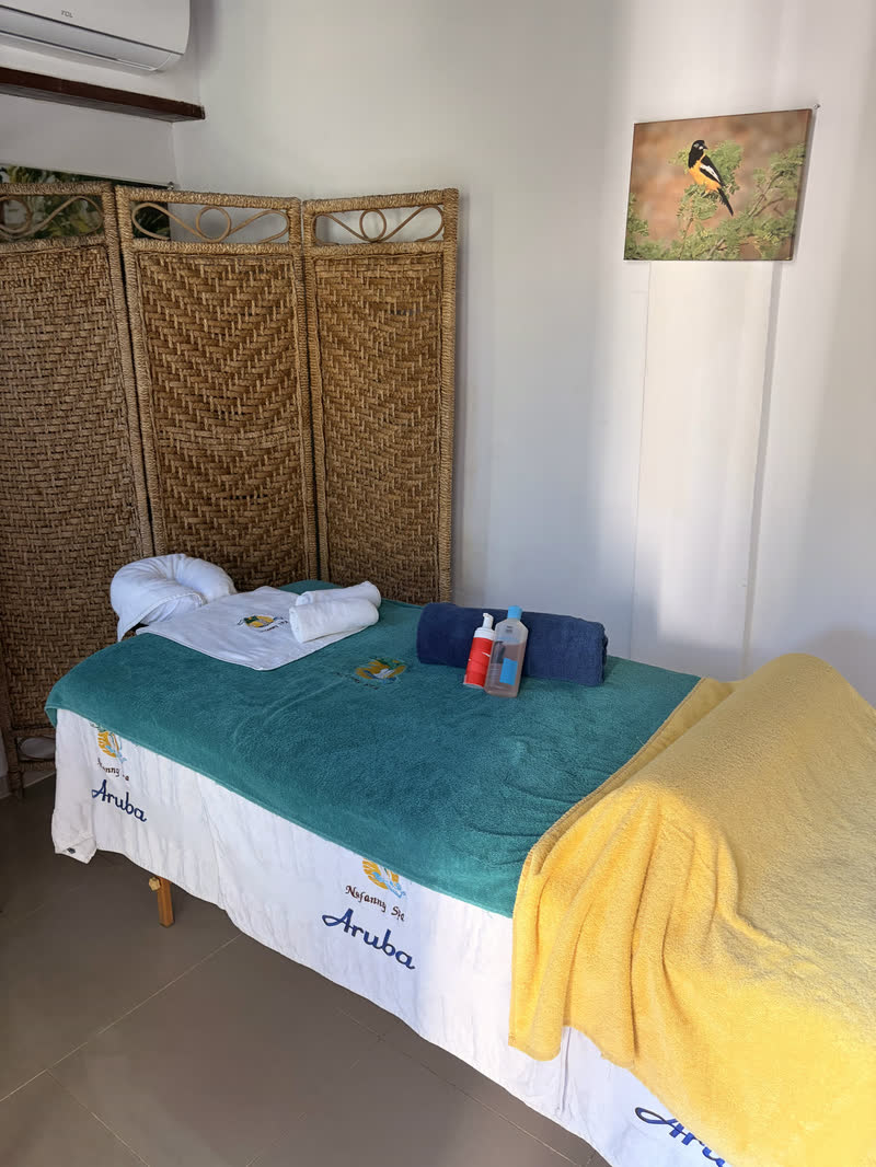 A massage table made up with teal and yellow towels in a treatment room, beside a woven folding screen