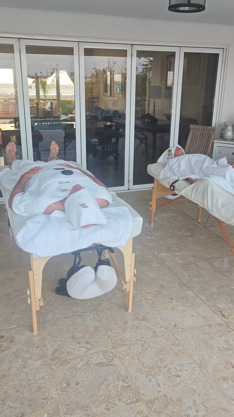 Two guests lying on portable massage tables on a covered terrace, under white towels, one with a massage stone resting on the towel; the glass doors behind them reflect the sea